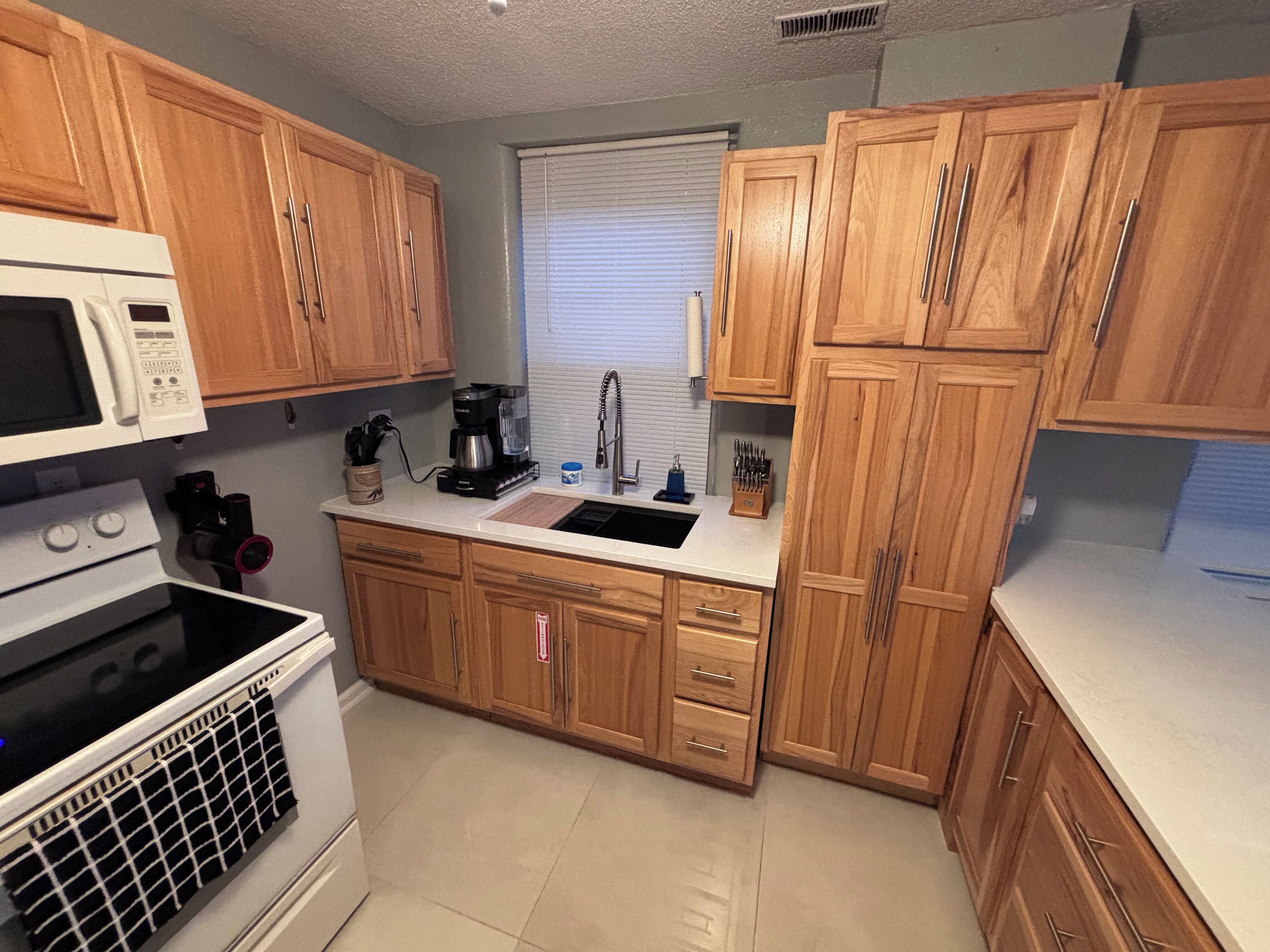 A kitchen with wooden cabinets, a white microwave, a stove, a sink, and a window with blinds.