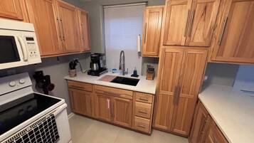 A kitchen with wooden cabinets, a white microwave, a stove, a sink, and a window with blinds.