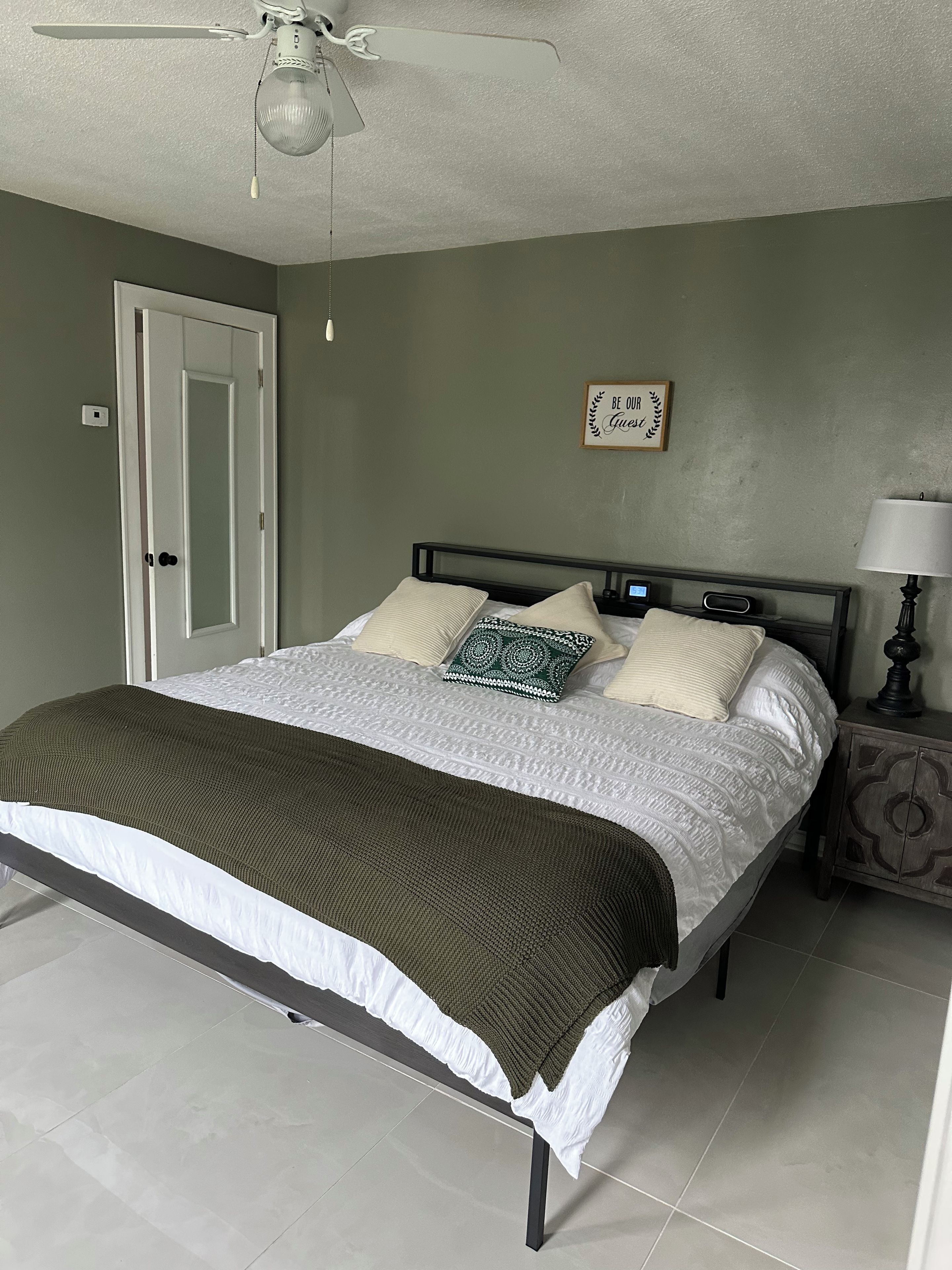 Ein Schlafzimmer mit einem Bett, einem Nachttisch mit Lampe, einem Deckenventilator, einer Tür und einer Wand mit einem Schild.