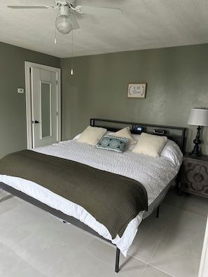 A bedroom with a bed, a bedside table with a lamp, a ceiling fan, a door, and a wall with a sign.