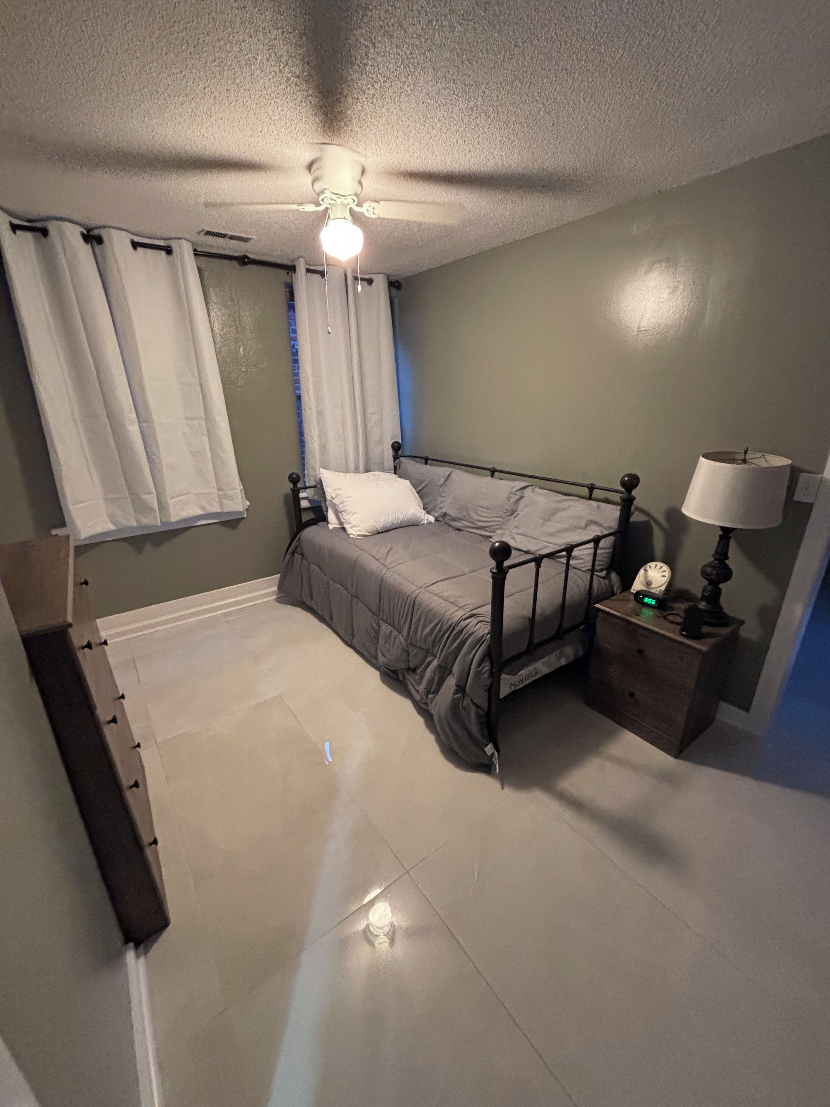 A bedroom with a bed, a nightstand with a lamp, a ceiling fan, and a window with curtains.