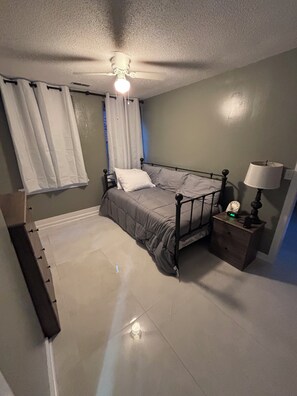 A bedroom with a bed, a nightstand with a lamp, a ceiling fan, and a window with curtains.