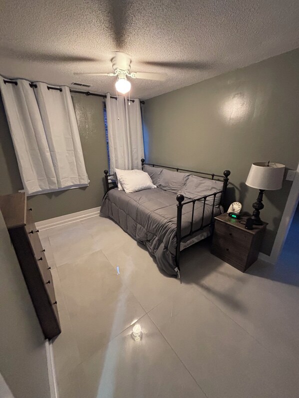Une chambre à coucher avec un lit, une table de chevet avec une lampe, un ventilateur de plafond et une fenêtre avec des rideaux.