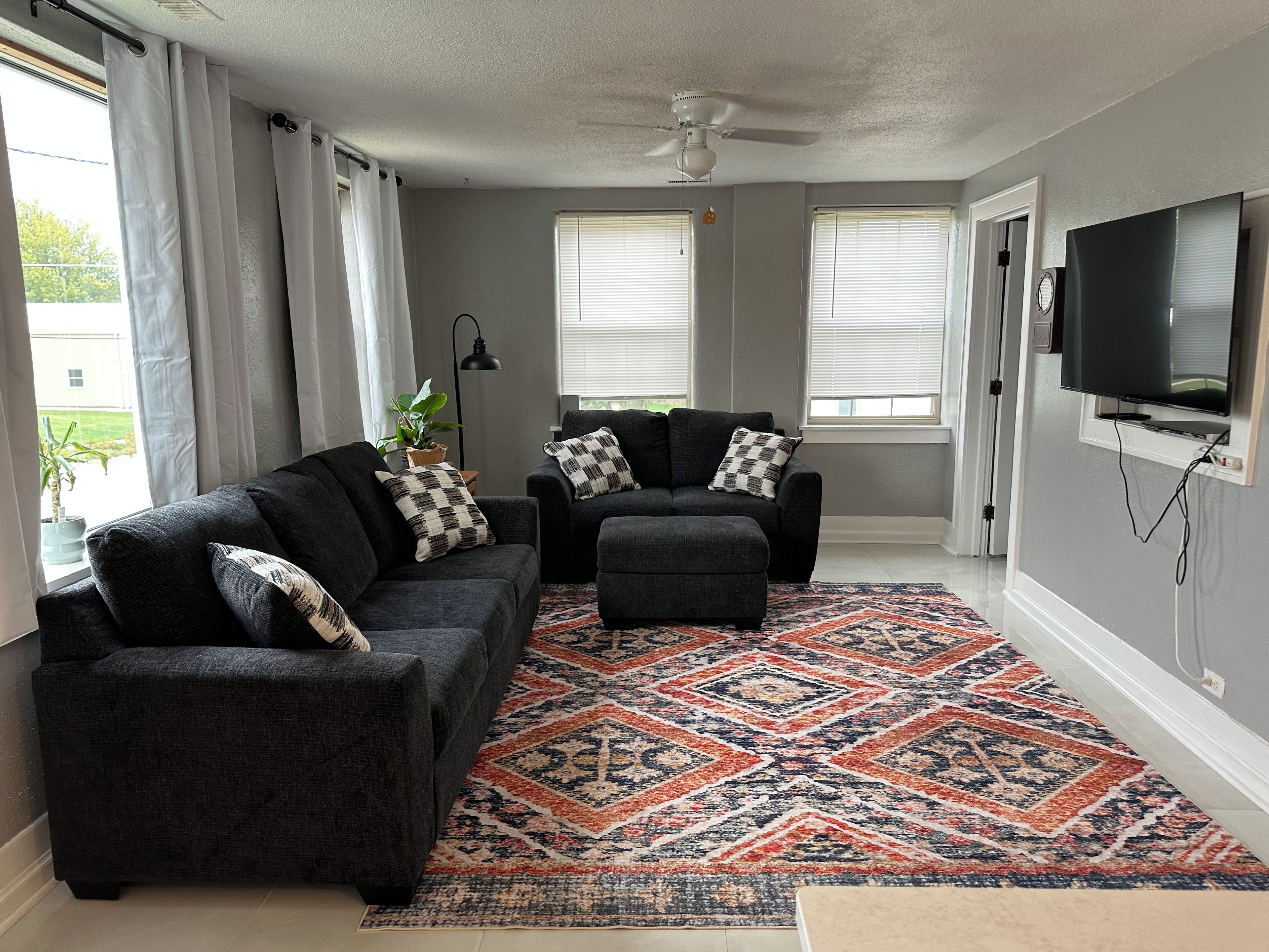 Un salon avec deux canapés, un tapis à motifs, un ventilateur de plafond et un téléviseur fixé au mur.