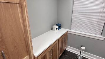 A bathroom with a wooden vanity, a white countertop, a toilet paper holder, and a window with blinds.