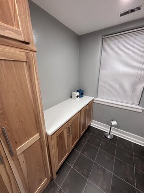 A bathroom with a wooden vanity, a white countertop, a toilet paper holder, and a window with blinds.