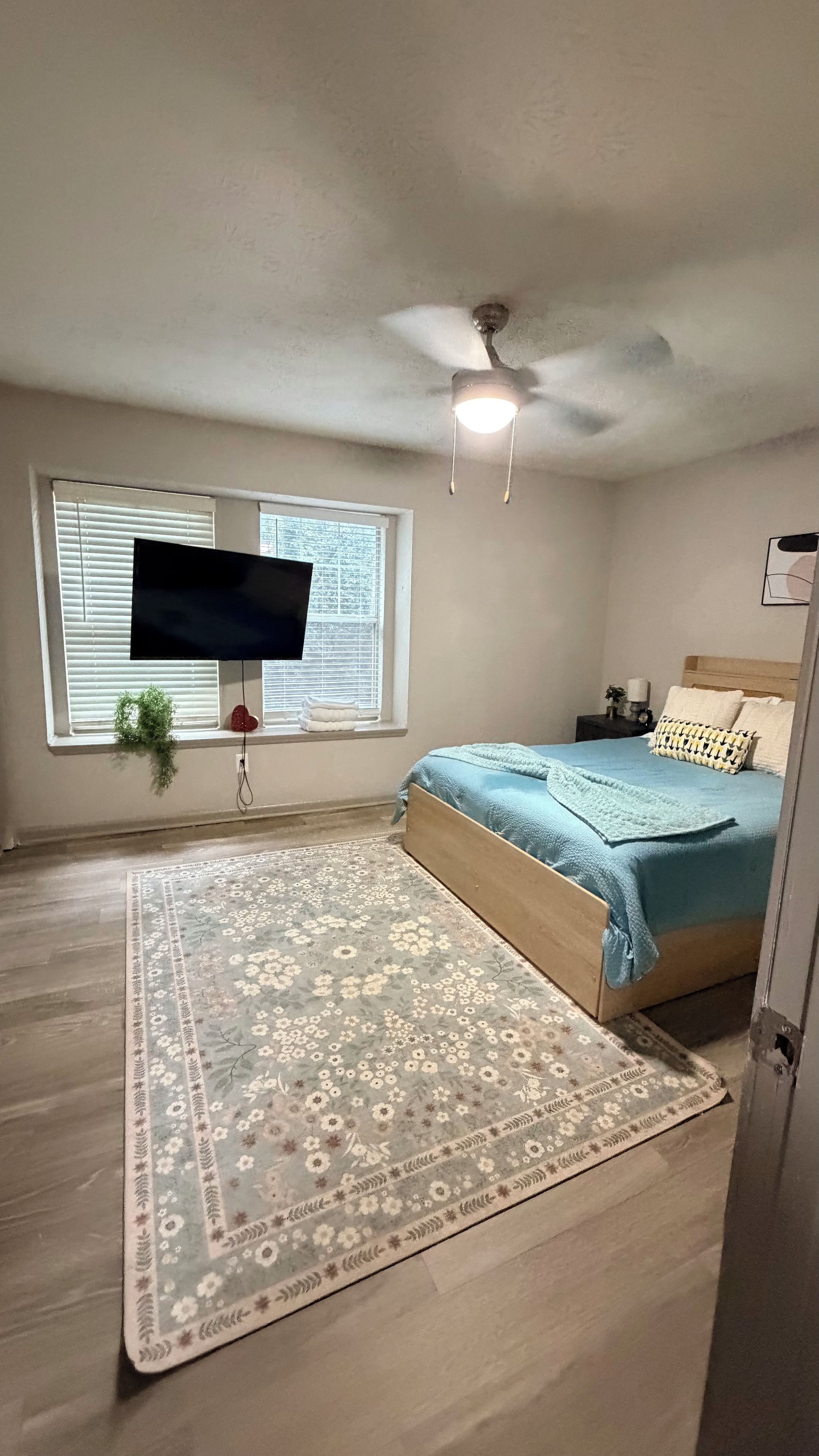 Un dormitorio con una cama, un televisor montado en la pared, un ventilador de techo, un ventanal con persianas y una alfombra estampada.