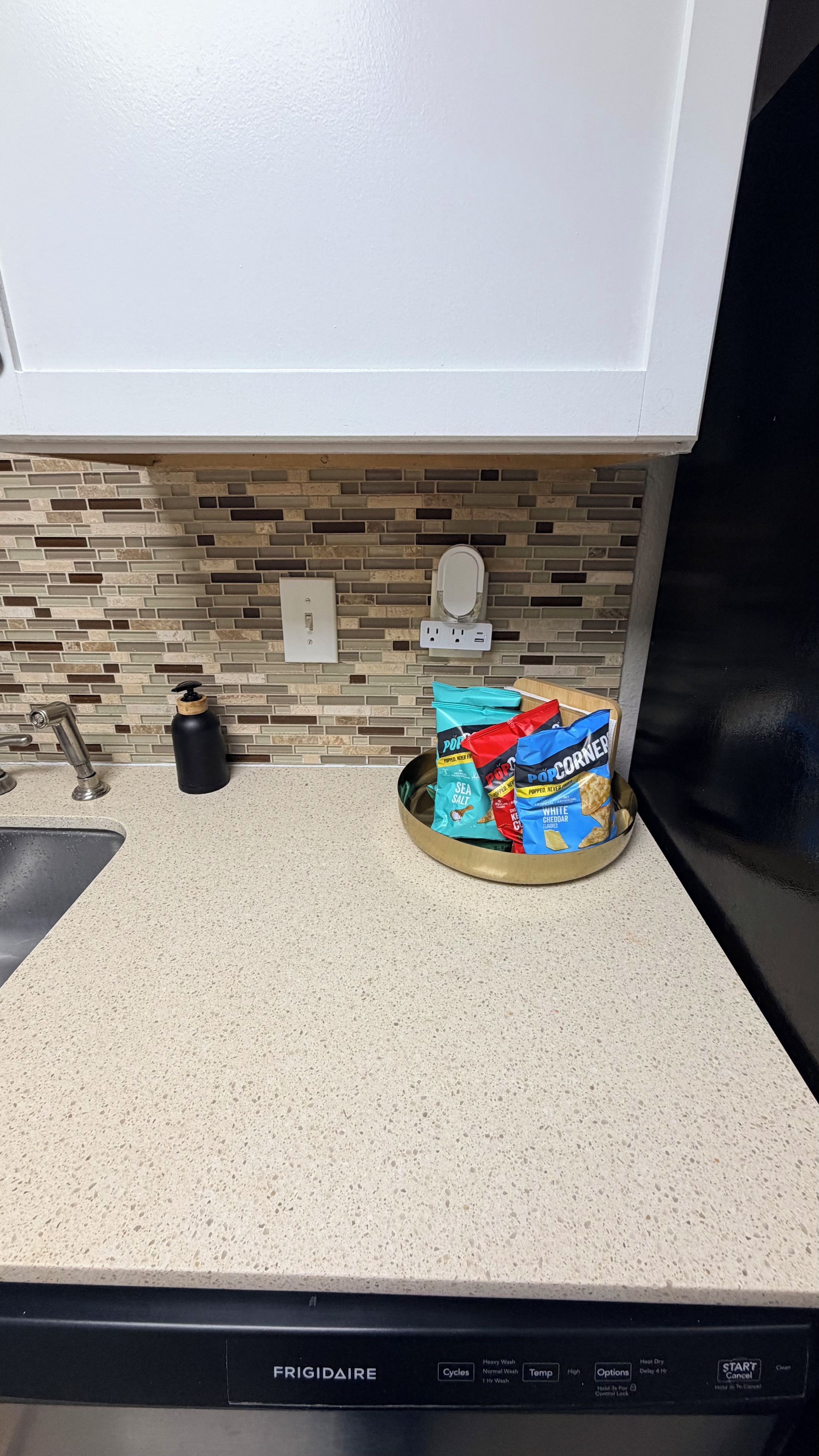 Una cocina con mostrador blanco, un lavamanos y un lavavajillas. Hay bolsas de condimento para palomitas en el mostrador.