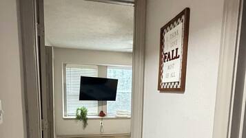 A hallway with a wooden floor leading to a room with a large window, a framed sign on the wall, and a plant by the window.