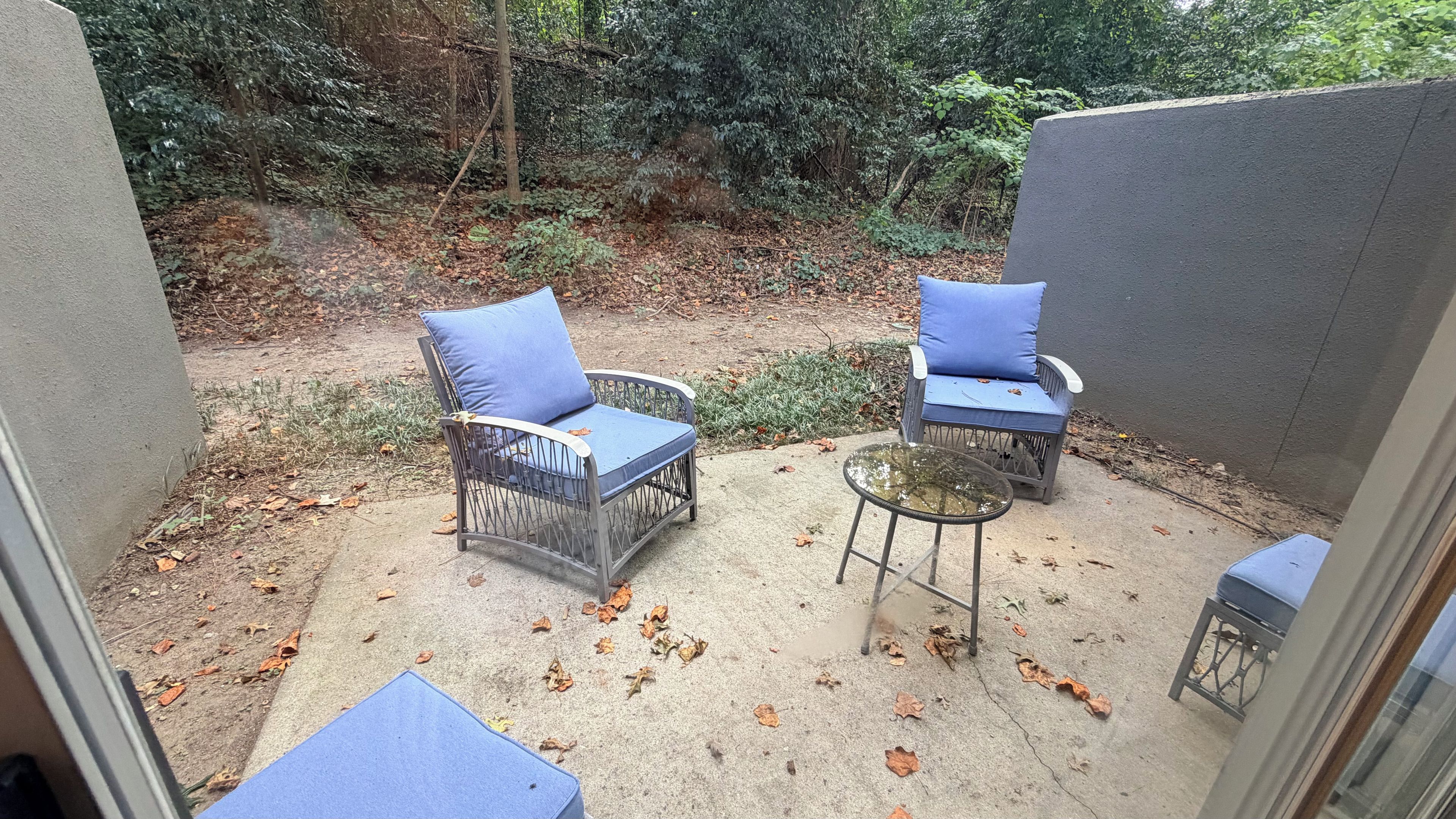 Un patio al aire libre con dos sillones de mimbre con cojines azules, una pequeña mesa redonda y un suelo de hormigón con hojas dispersas.