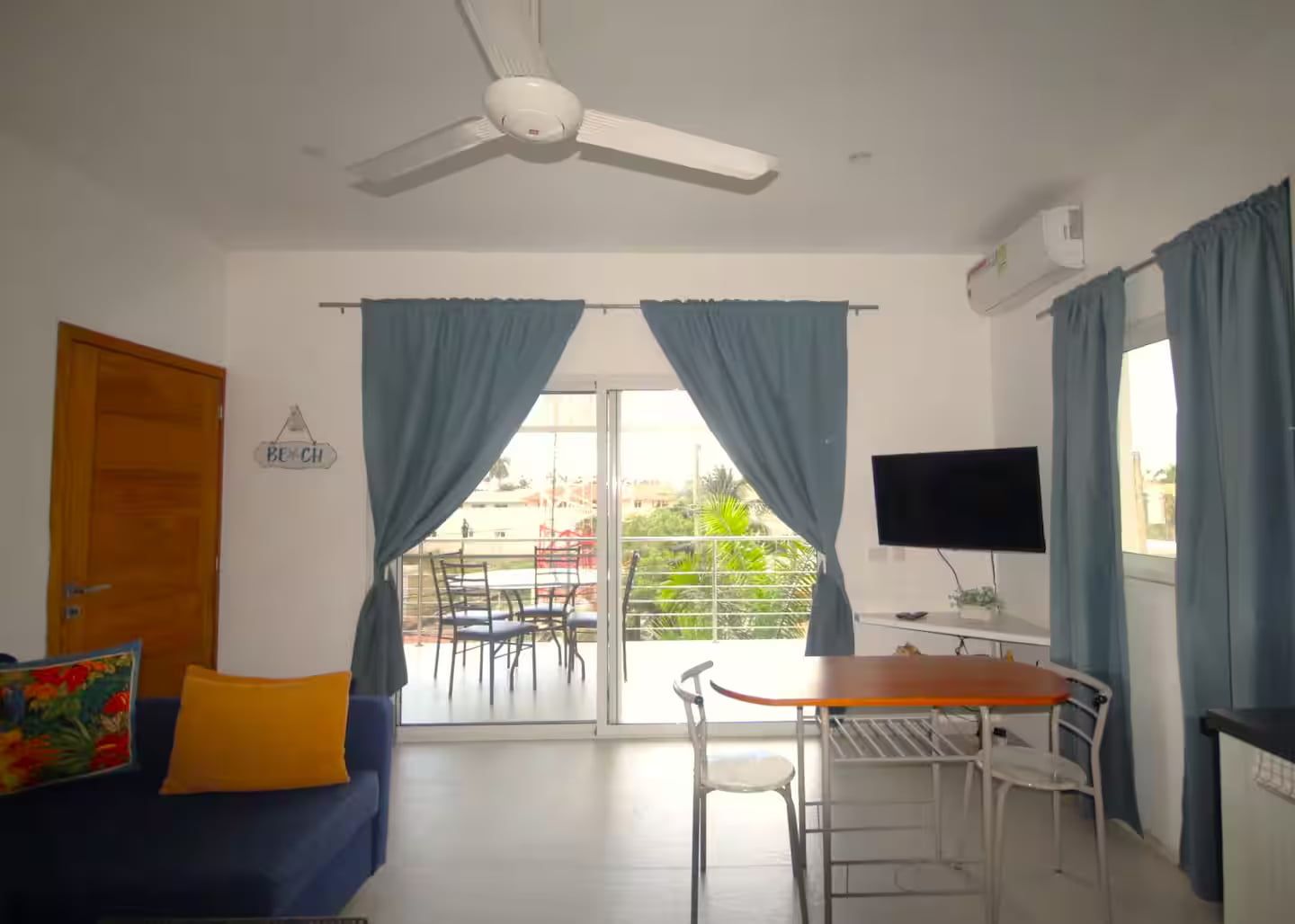 A room with a ceiling fan, a TV, a dining table with chairs, a sofa with a cushion, and a balcony with chairs and curtains.