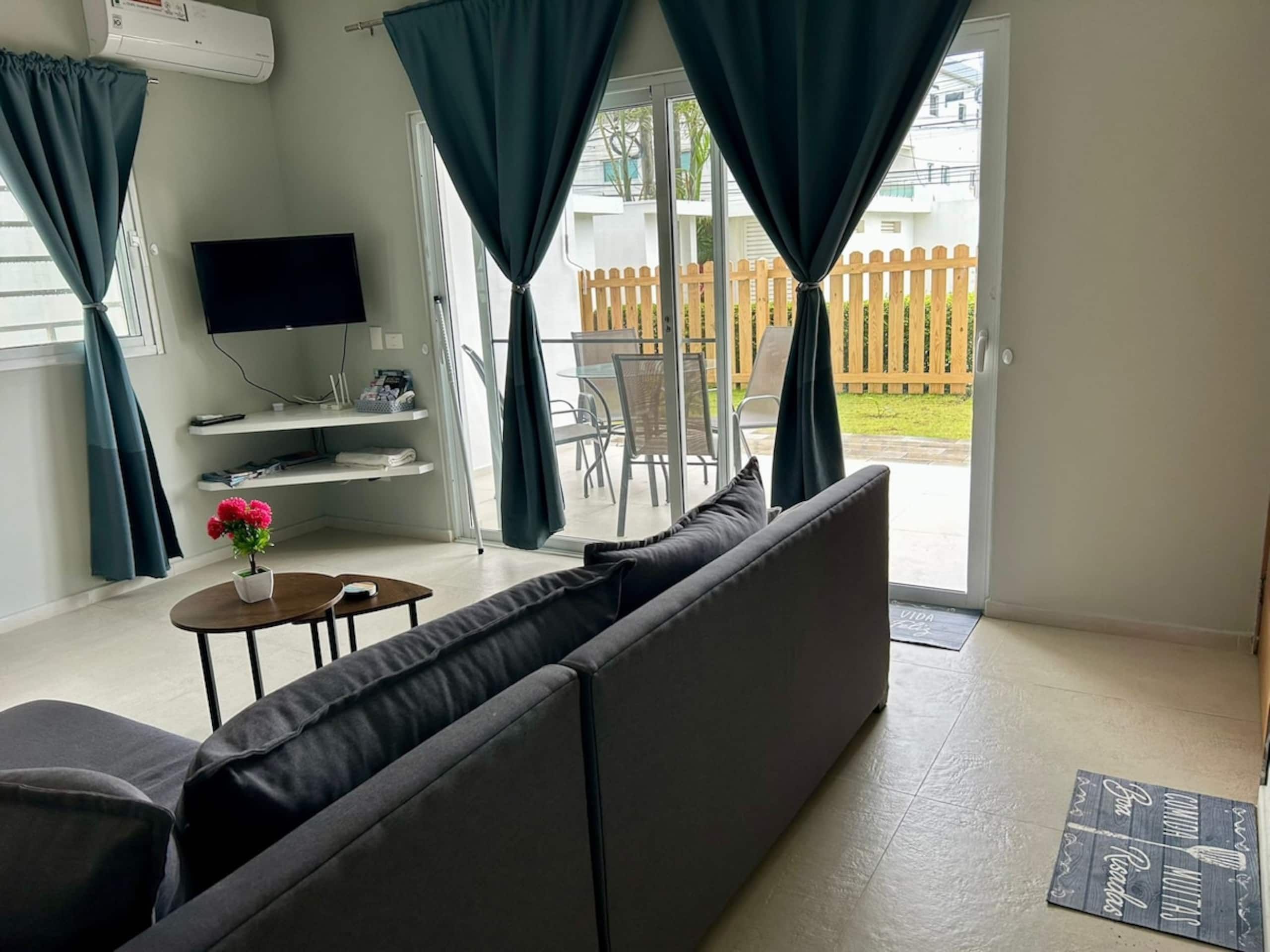 Ein Wohnzimmer mit einer Couch, einem Fernseher, einem kleinen Tisch mit einer Pflanze und Blick ins Freie.