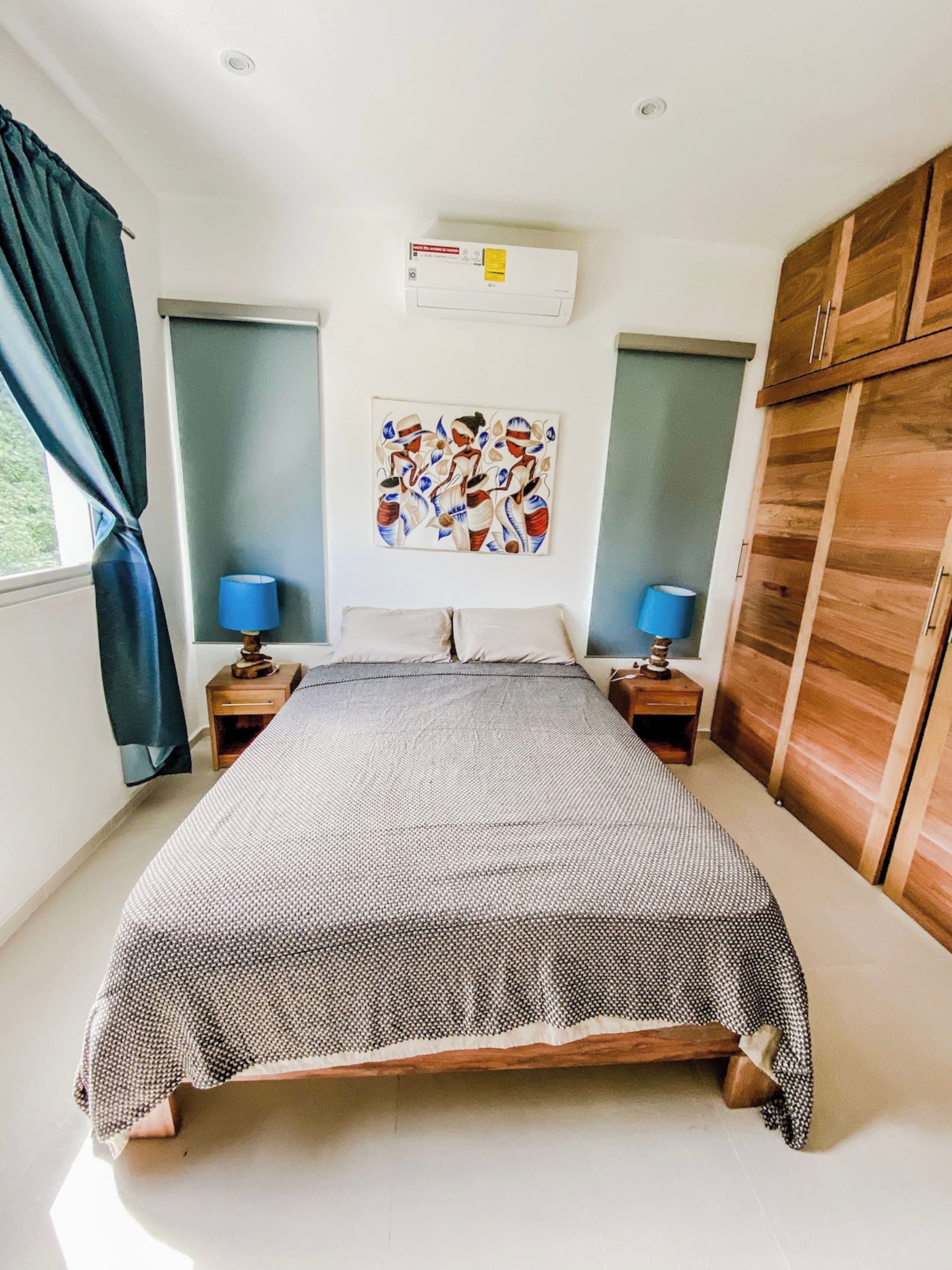 Une chambre à coucher avec un grand lit, deux tables de chevet avec des lampes, un climatiseur fixé au mur et une armoire en bois.