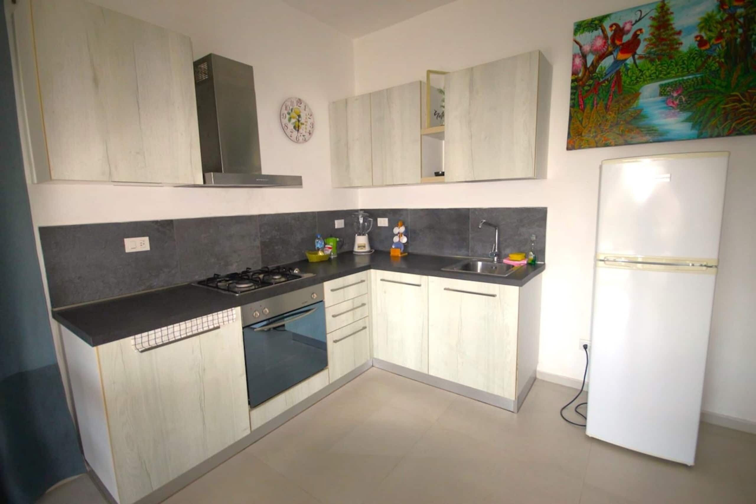 A modern kitchen with a gas stove, oven, and sink. There is a refrigerator and a clock on the wall.
