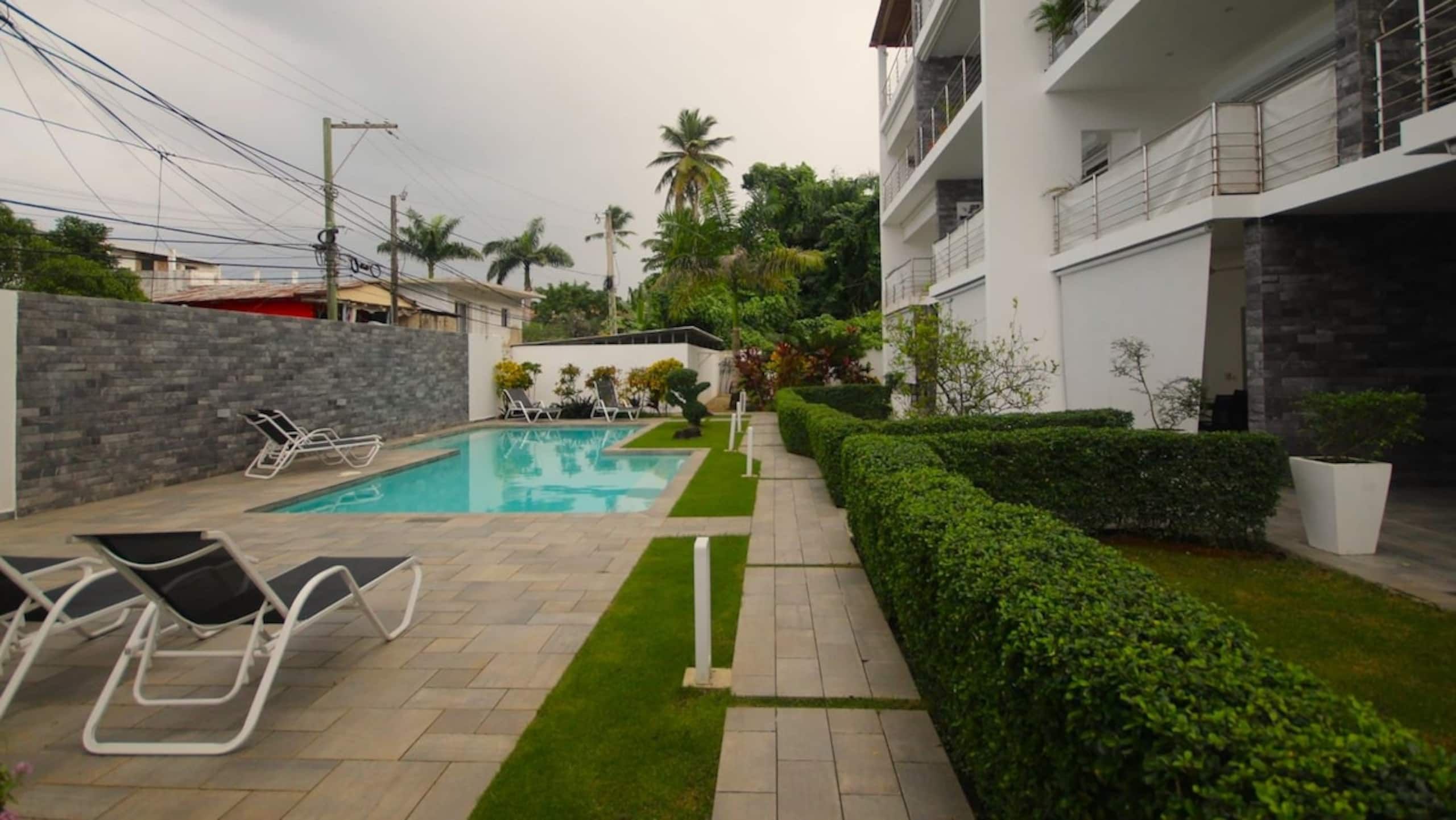 A swimming pool with lounge chairs, a well-maintained lawn, and a modern building with balconies.