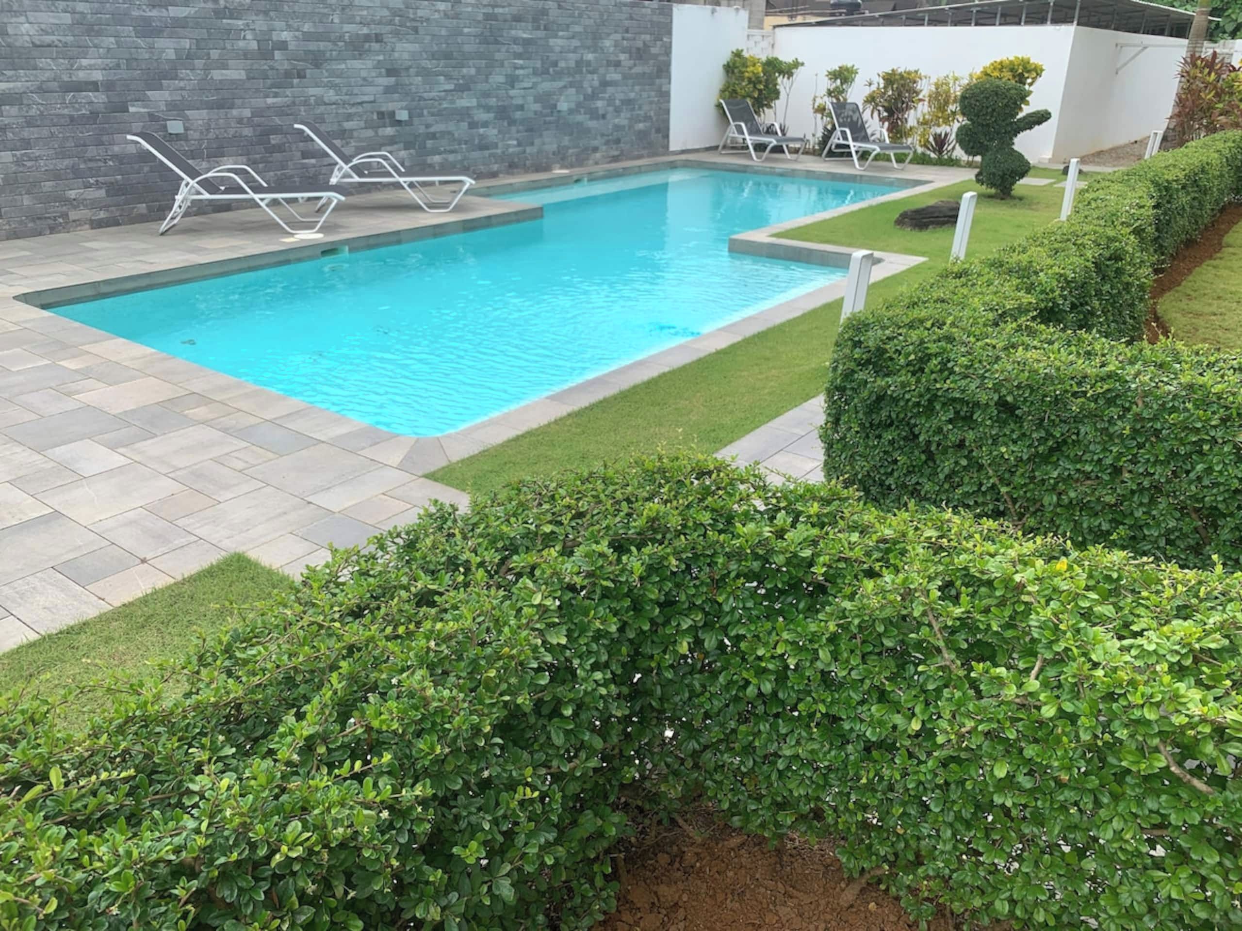 Una piscina con solárium de baldosas, rodeada de setos cuidados y un muro de piedra.