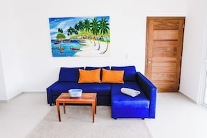 Una habitación con un sofá azul, dos cojines naranjas, una mesa de centro de madera y un cuadro con una playa.