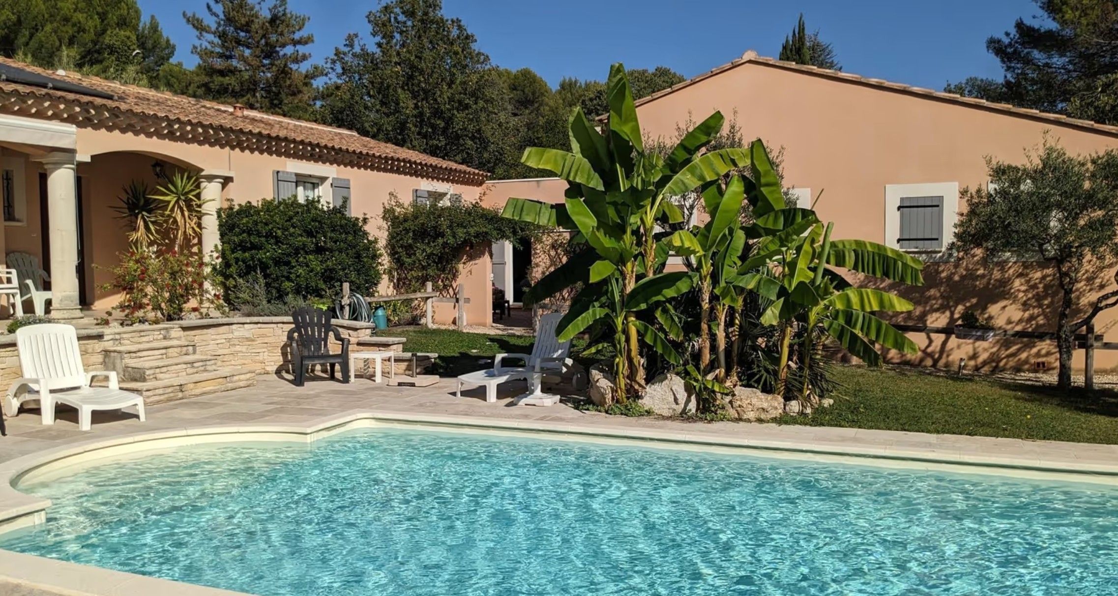 Une piscine dotée d’une terrasse carrelée, entourée d’un jardin planté de bananiers et bordée d’un bâtiment dont le toit est également recouvert de tuiles.