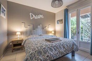 A bedroom with a bed, two bedside tables, a wall-mounted light fixture, and a view of greenery outside.