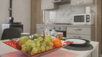 Uma mesa de jantar com uma tigela de frutas contendo uvas, uma laranja e uma maçã verde. Ao fundo, há uma área de cozinha com micro-ondas e armários.