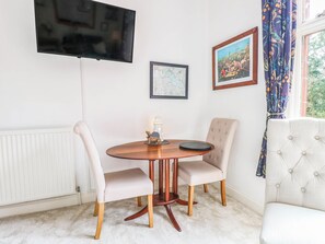 A dining area with a round table, two chairs, a television mounted on the wall, and a window with curtains. - Heritage Hideaway (Norwich)