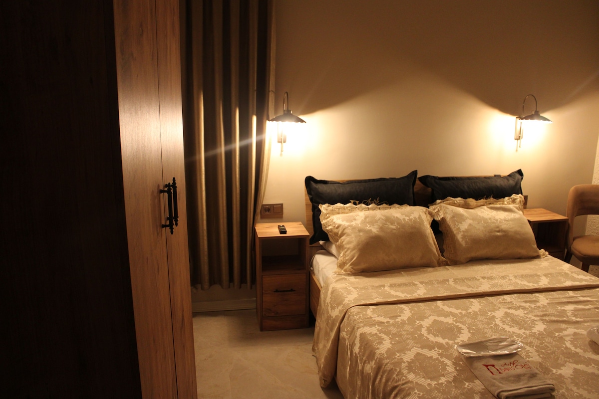 Una habitación de hotel con una cama, mesitas de noche, un sillón y una cortina.
