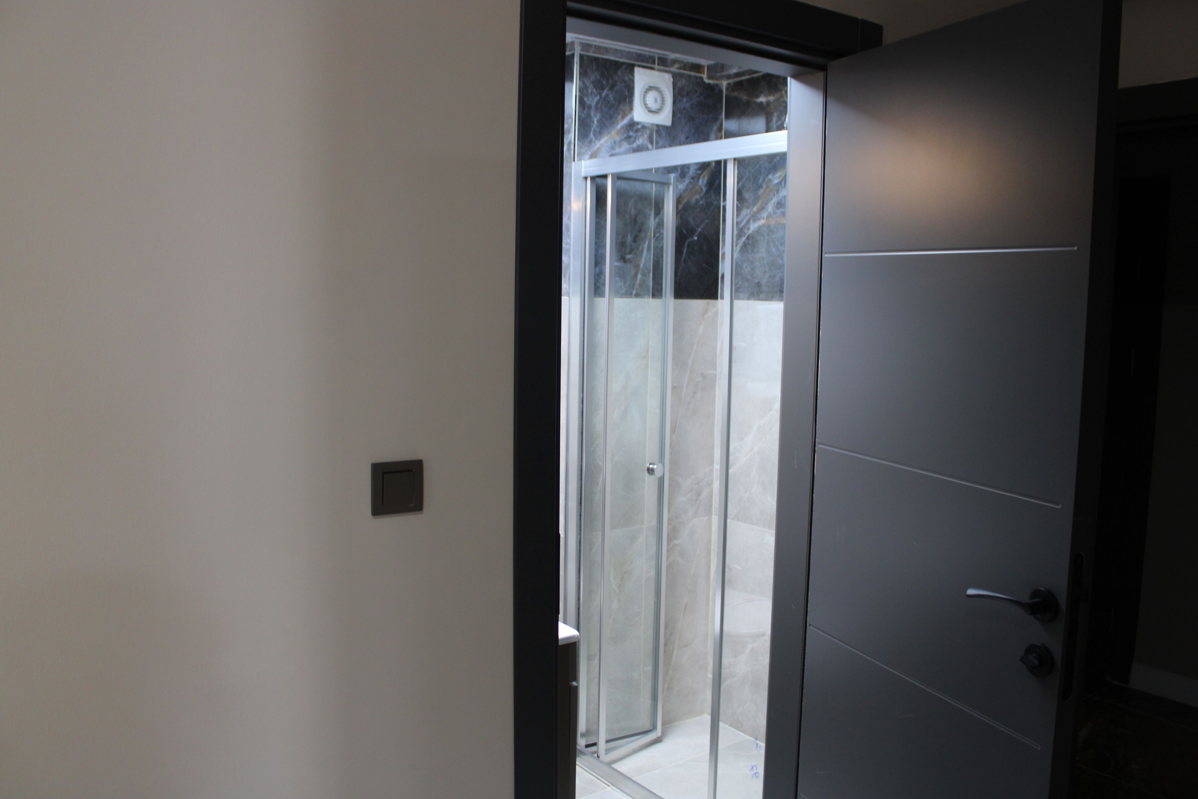 A modern bathroom with a glass shower enclosure, a dark door, and a visible light switch on the wall.