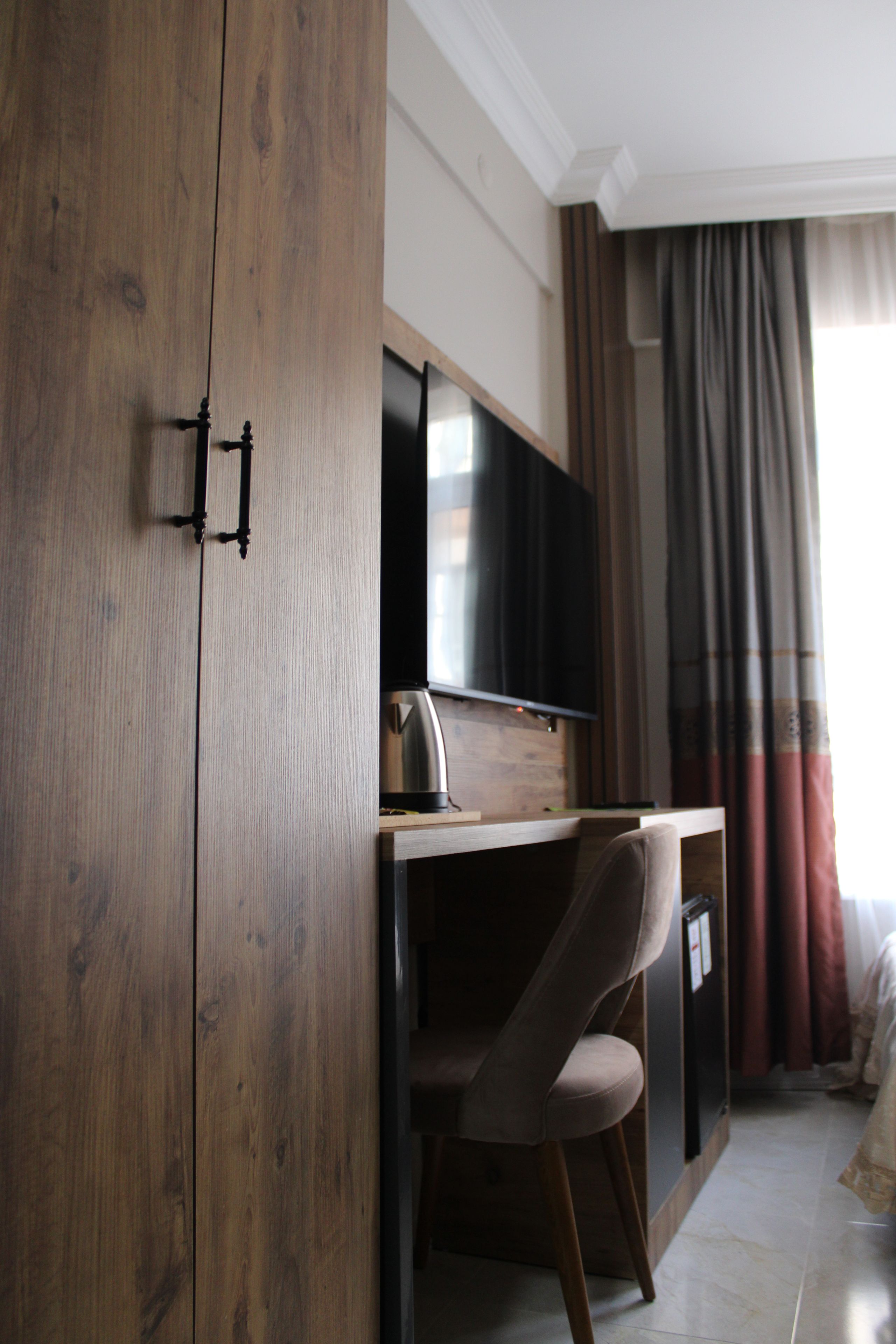 Una habitación con un armario de madera, un escritorio con una silla, un televisor de pantalla plana y una ventana con cortinas.