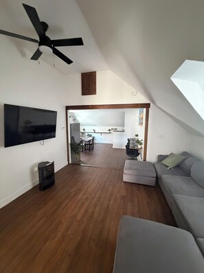 Ein großzügiger Wohnbereich mit Deckenventilator, einem Flachbildfernseher, einem grauen Sofa und einem Essbereich im Hintergrund.
