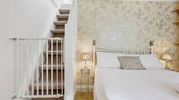 A bedroom with a bed, a nightstand with a lamp, a white railing on a staircase, and a floral wallpaper pattern.