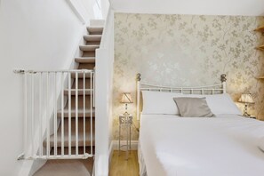 A bedroom with a bed, a nightstand with a lamp, a white railing on a staircase, and a floral wallpaper pattern.