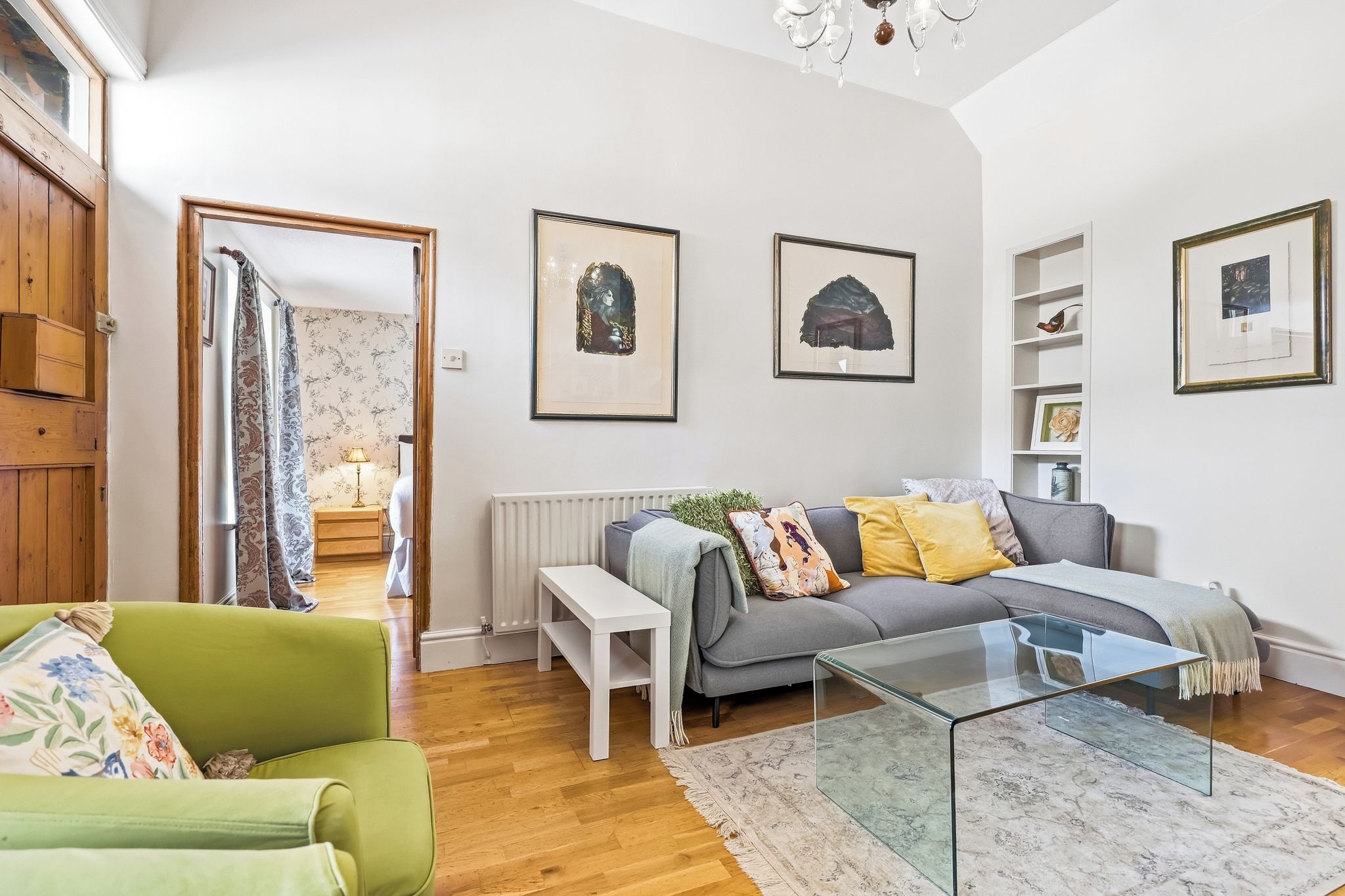 Un salon comprenant un canapé, une table basse en verre, un fauteuil vert et un radiateur. Des œuvres d’art encadrées ornent le mur, et une porte en bois donne accès à une autre pièce.