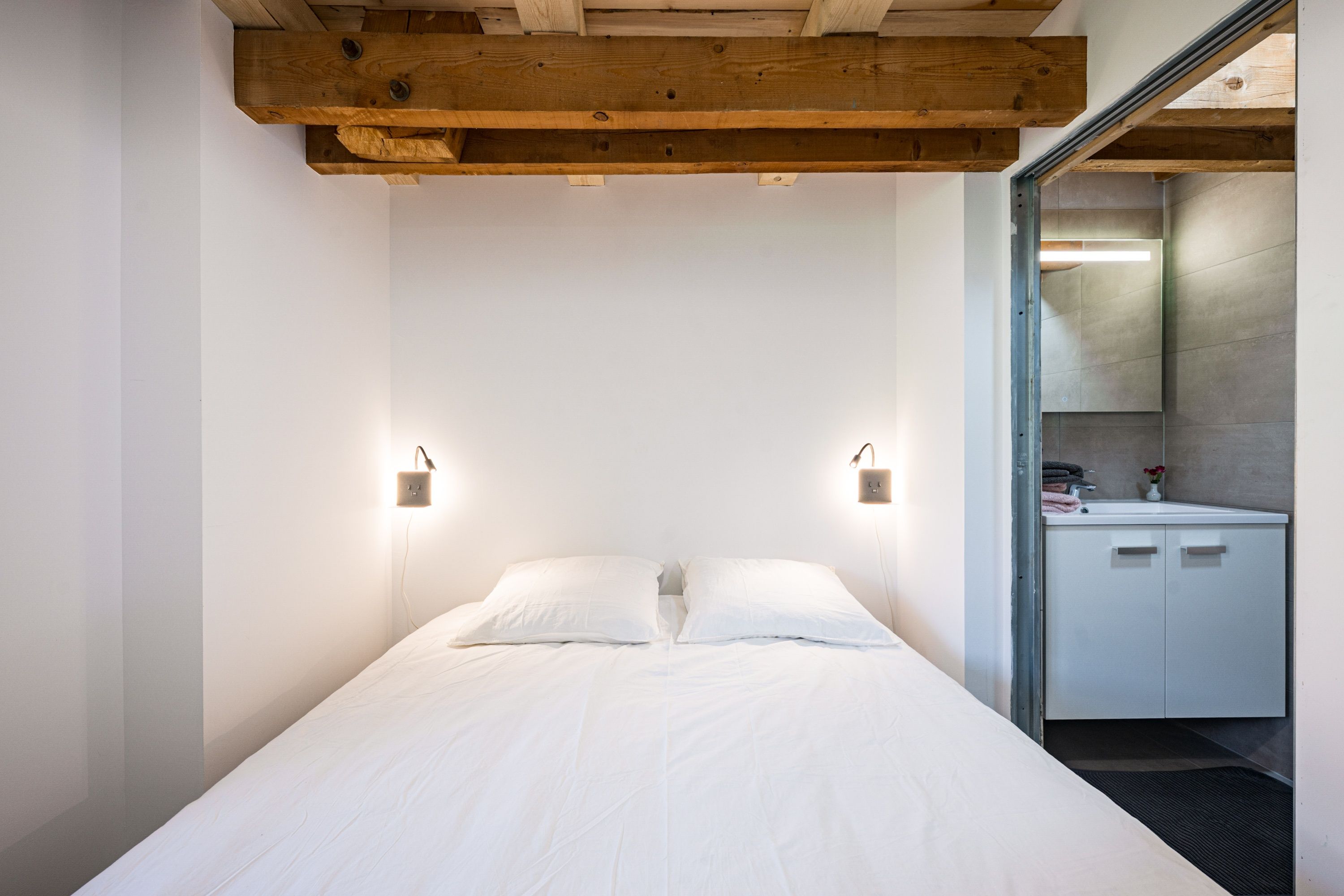 Quarto com cama, duas luminárias de cabeceira, uma viga de madeira no teto e um banheiro visível por uma porta aberta.