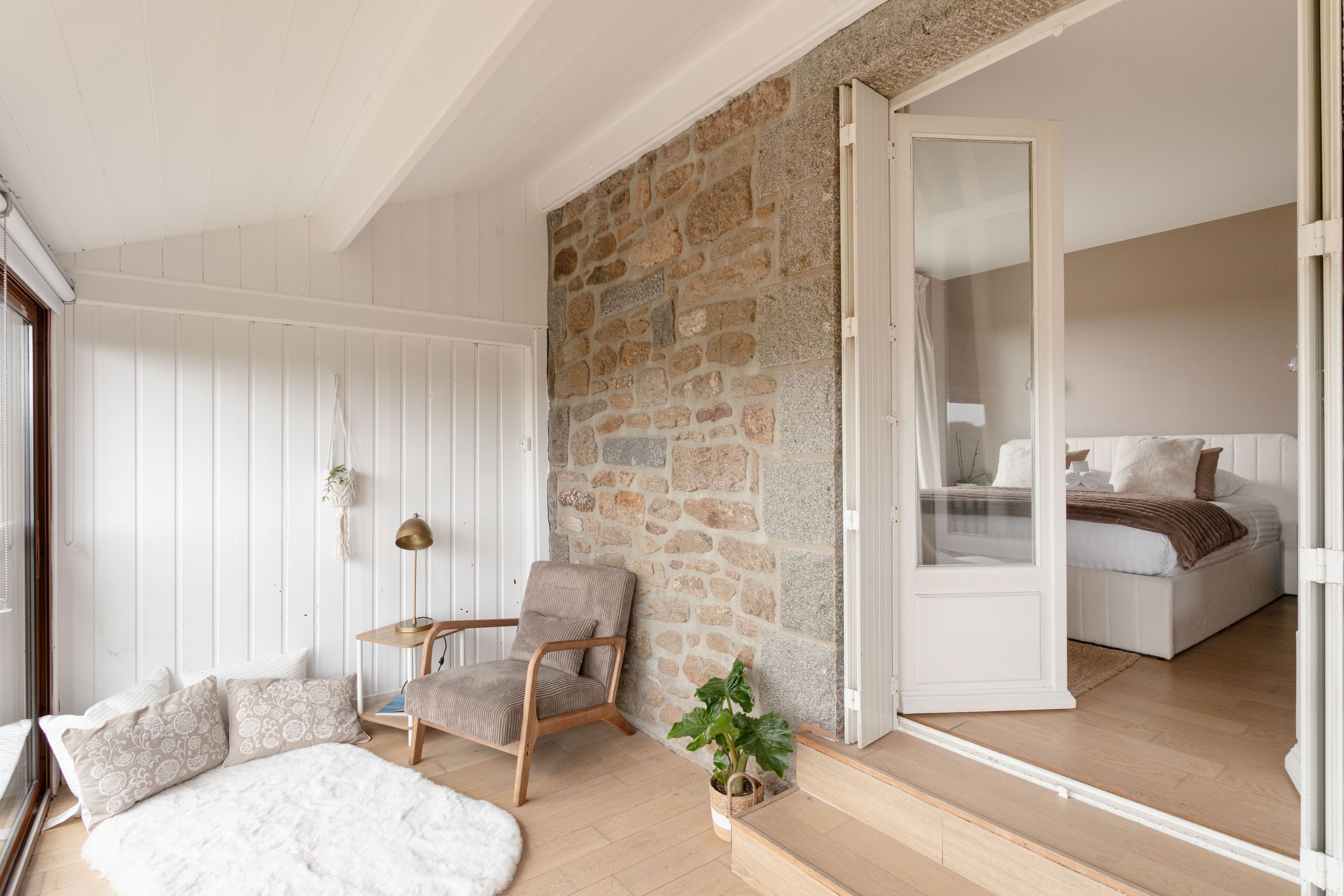 A cozy room with a stone wall, a bed with white bedding, a chair, and a small table with a lamp.