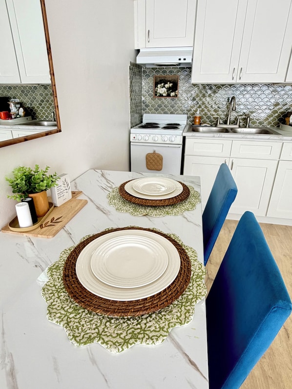 Een keuken met witte kasten, een fornuis en een gootsteen. Op een tafel liggen twee borden met een bruin-groene placemat.