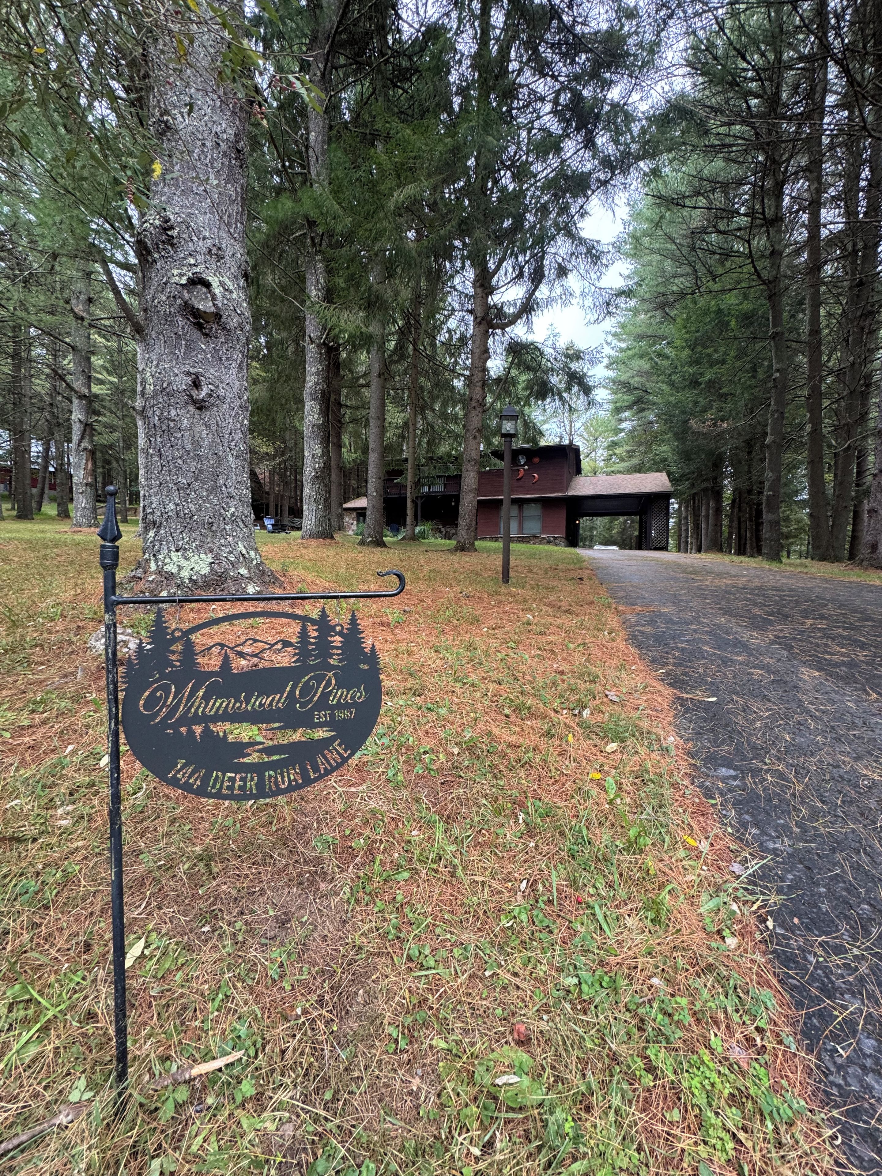 Placa para 'Whimsical Pines' com design rústico, um caminho pavimentado e um edifício ao fundo.
