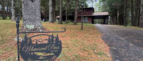 Placa para 'Whimsical Pines' com design rústico, um caminho pavimentado e um edifício ao fundo.