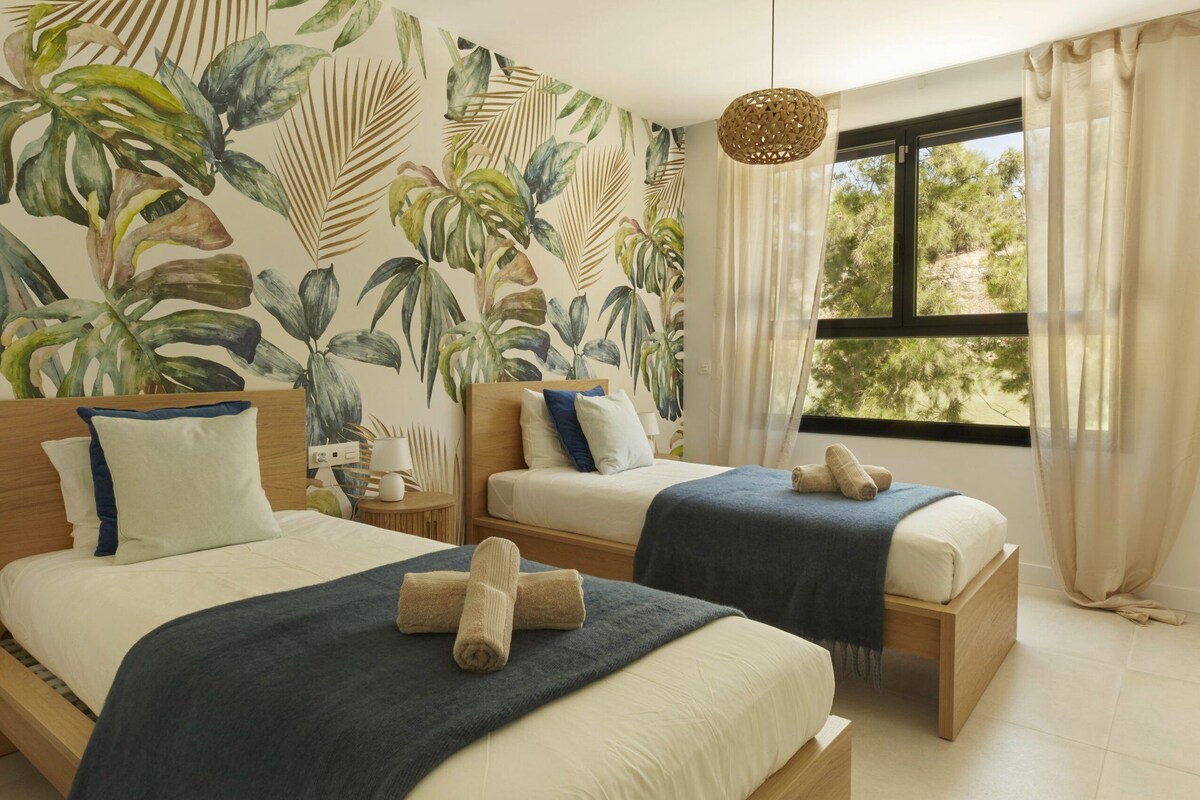 Ein Schlafzimmer mit zwei Betten, tropischen Blättermotiven an der Wand, einer Hängeleuchte und einem Fenster mit Vorhängen.