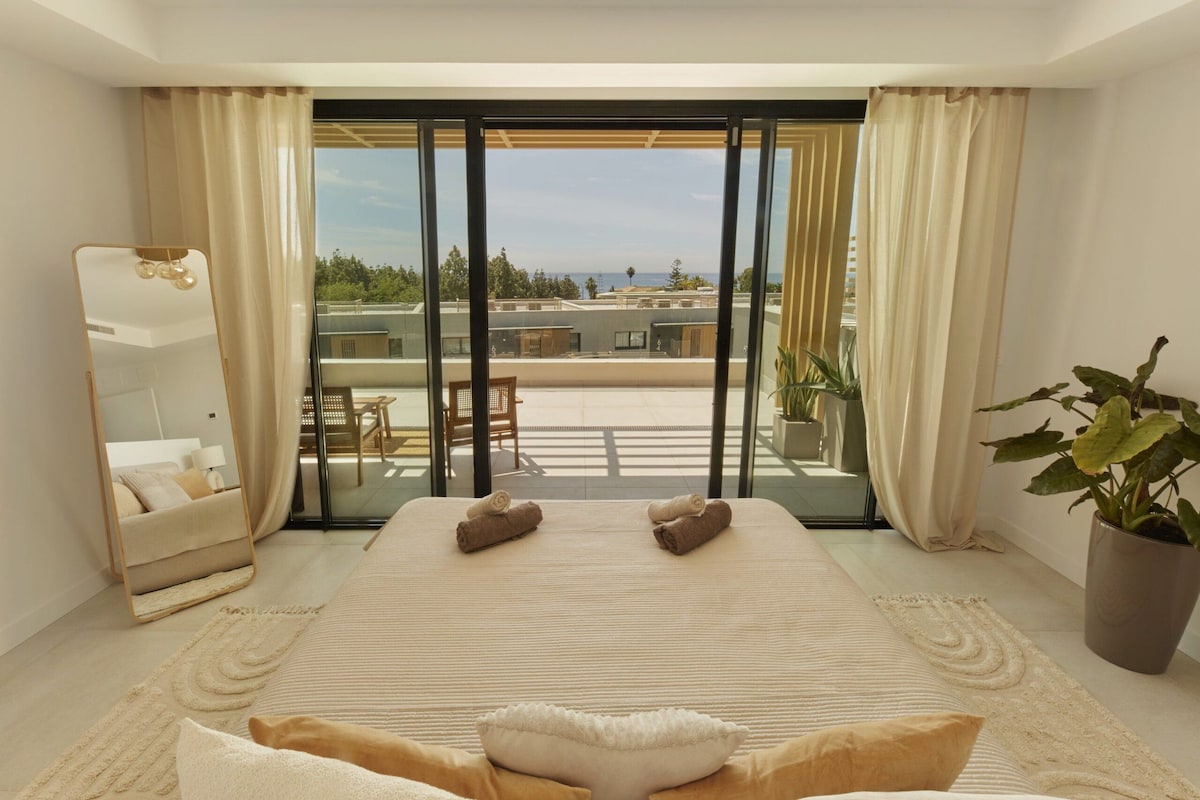 A bedroom with a large bed, a sitting area, a balcony with a view, and a potted plant.