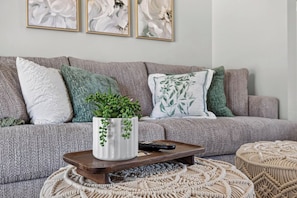 A living room with a grey sofa, green and white patterned cushions, a white vase with green plants, a remote control, and a woven ottoman. - Spacious 3BR Home with Full Kitchen and Carport (Abilene)