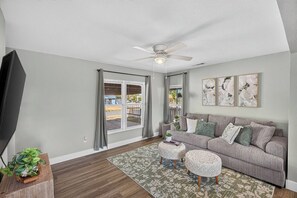 A living room with a grey sofa, a flat-screen TV, a ceiling fan, and a large window with curtains. - Spacious 3BR Home with Full Kitchen and Carport (Abilene)