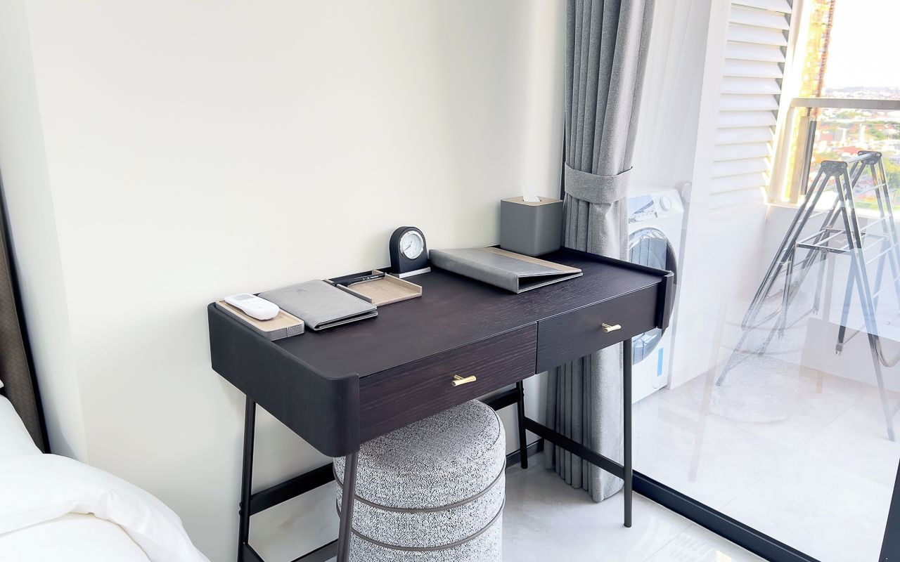 Un dormitorio moderno con un escritorio de madera oscura, una mesita de noche, una lámpara y una ventana con cortinas.