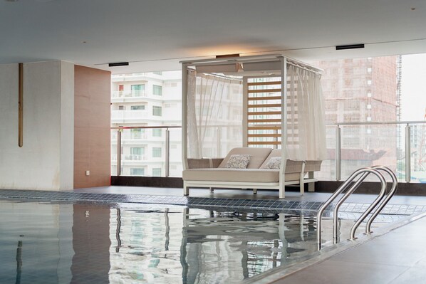 Un área de piscina interior moderna con piso de vidrio, un sofá blanco con cojines estampados y vista a un paisaje urbano a través de amplias ventanas.