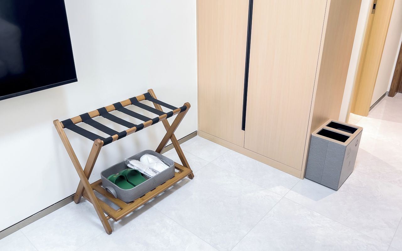 A room with a wooden folding chair, a small storage bin, a trash bin, and a television mounted on the wall.