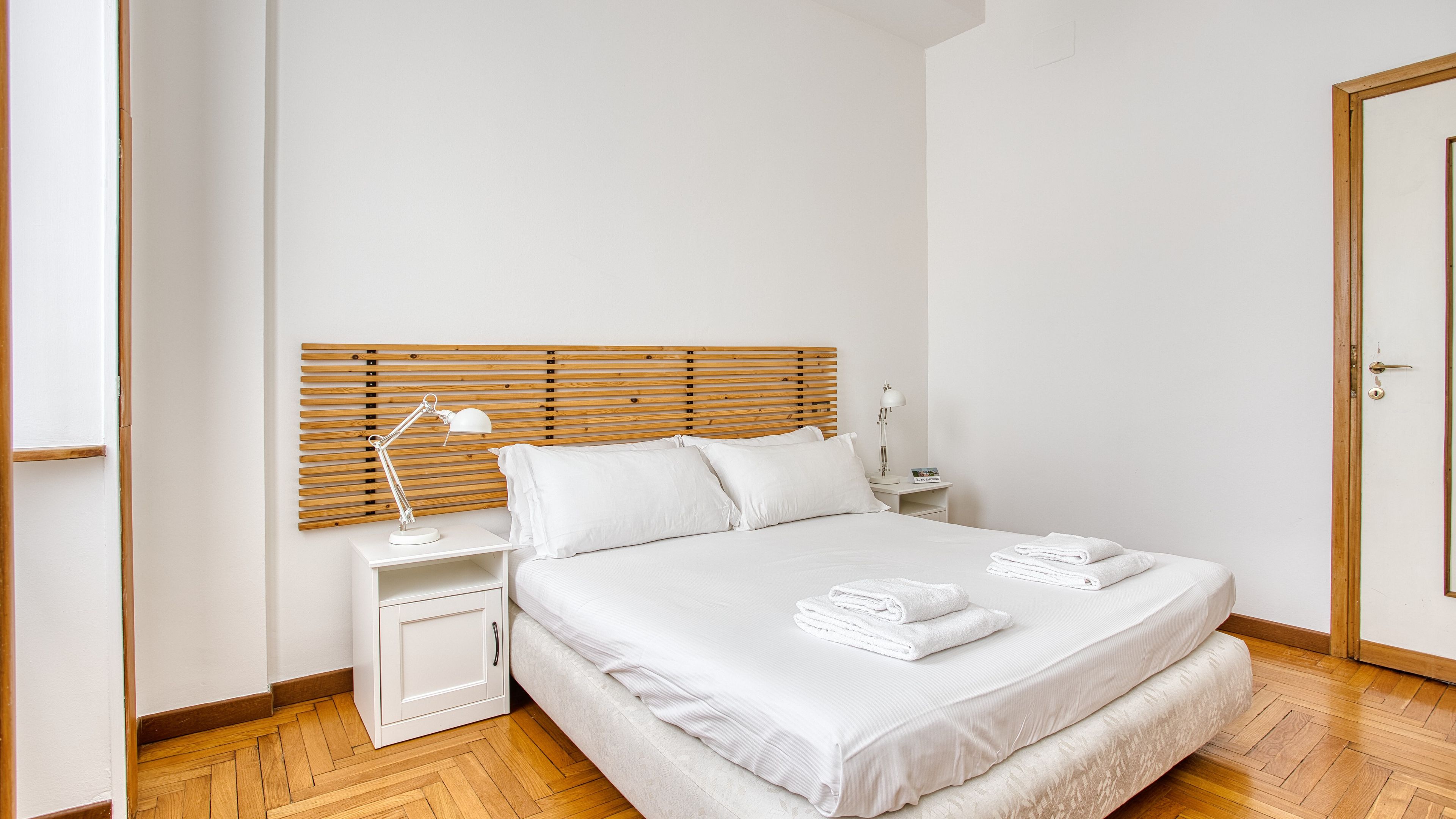 Quarto com cabeceira de madeira, cama branca, duas mesas de cabeceira, abajur branco e toalhas dobradas com cuidado.