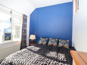 A bedroom with a bed, a window with curtains, a bedside table with a lamp, and a wall with a blue accent. - Angels Place (Yelverton)