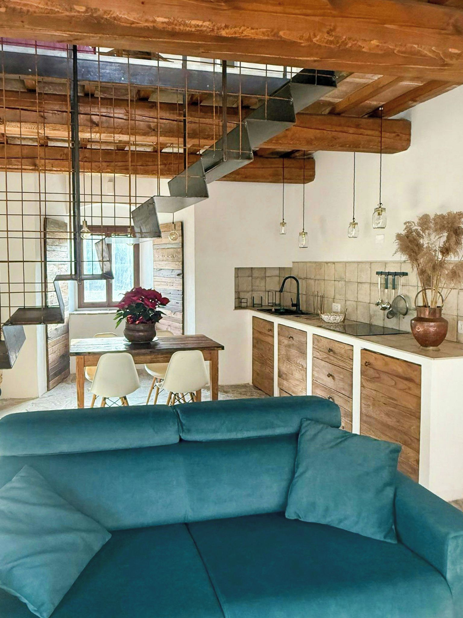 Un intérieur moderne avec un canapé bleu, des poutres en bois au plafond, une cuisine équipée d’un évier et des luminaires suspendus.