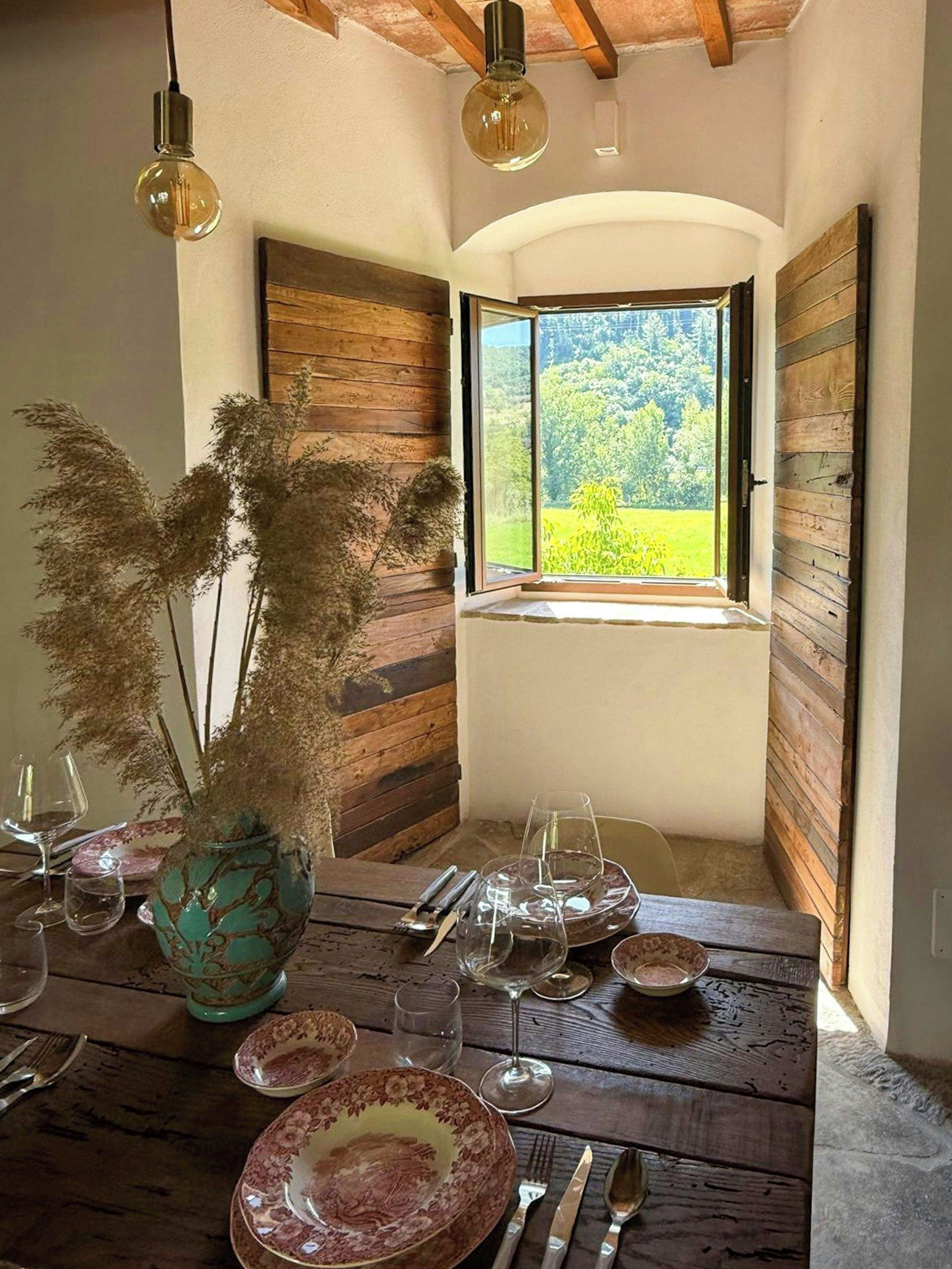Zona pranzo con tavolo in legno rustico, un vaso con piante essiccate e una finestra vista verde.