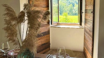 Zona pranzo con tavolo in legno rustico, un vaso con piante essiccate e una finestra vista verde.