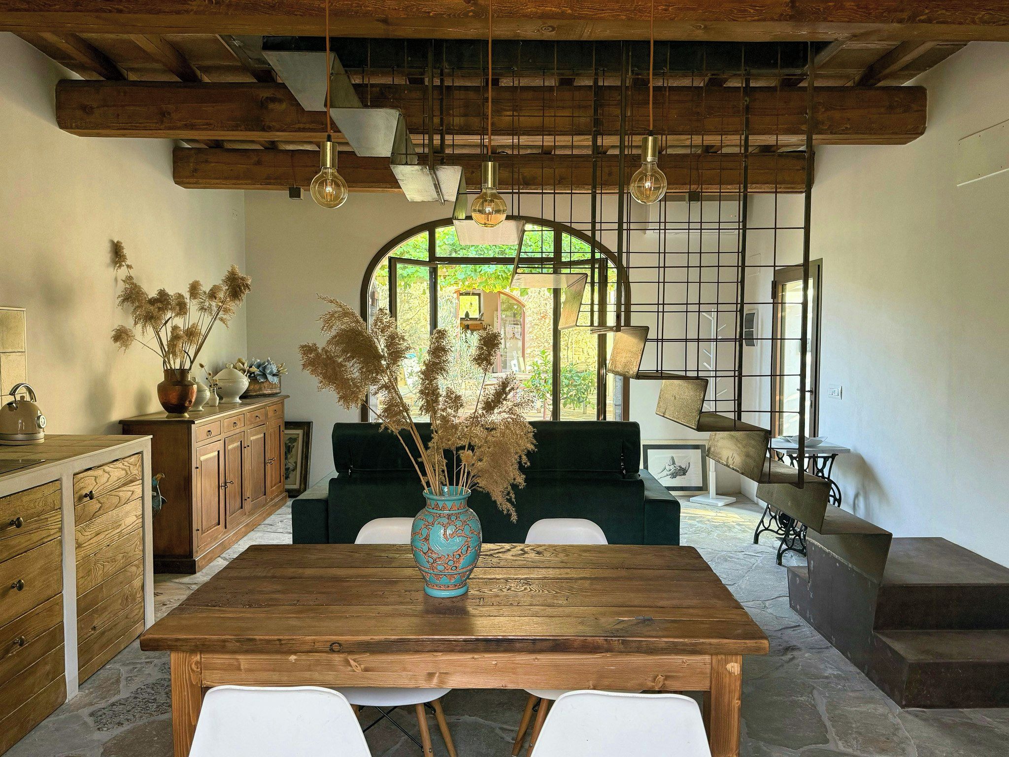 Un interno rustico con un tavolo da pranzo in legno, un vaso con piante essiccate e una vista attraverso una finestra con inferriata metallica.